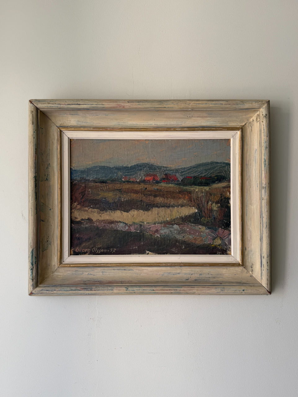 A framed vintage landscape oil painting. The scene depicts a field with distant hills and small red houses. Available at Hue.