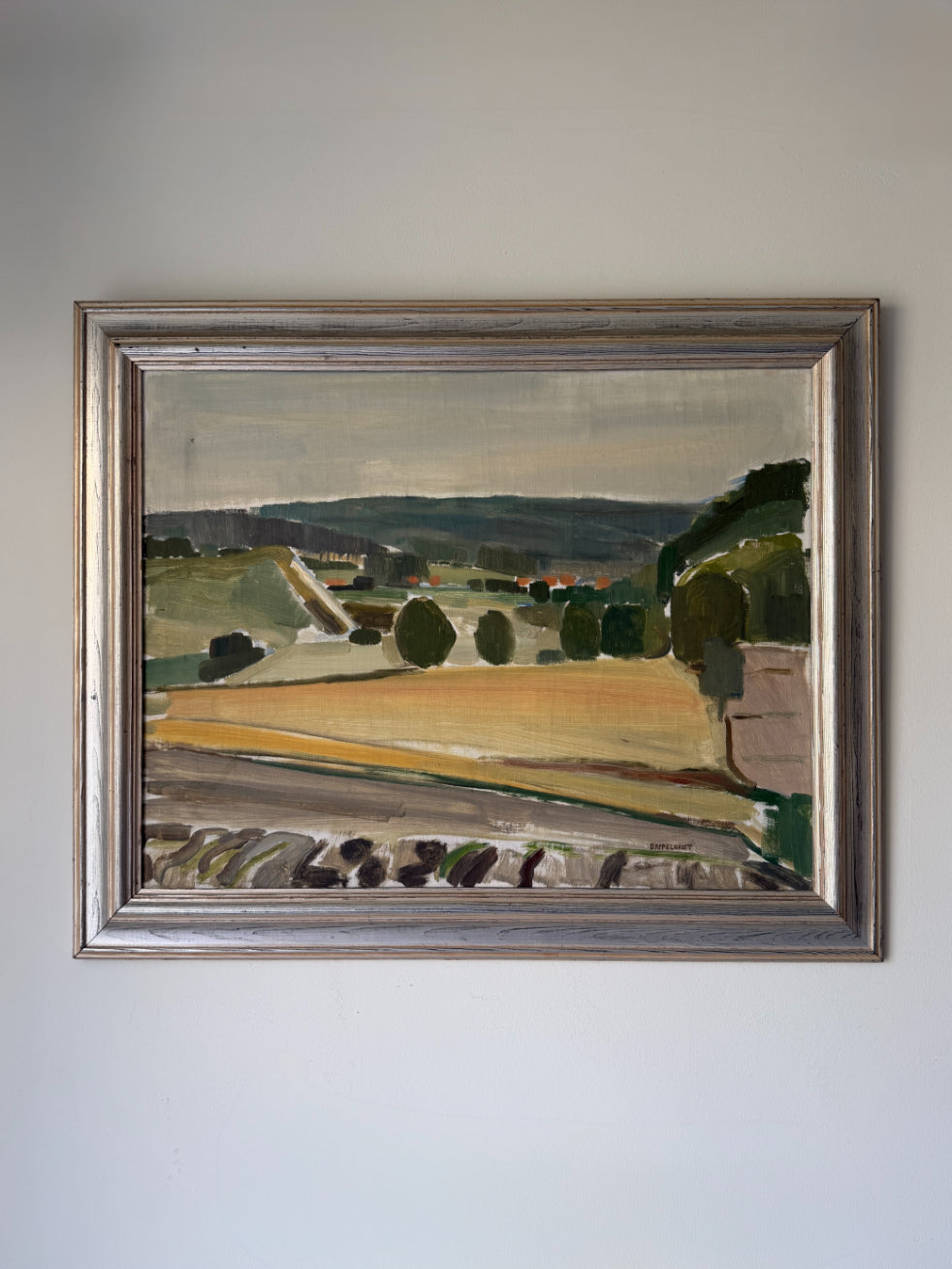 A framed vintage landscape oil painting featuring green hills, trees, and a golden field, housed in a distressed silver frame. Available at Hue. 