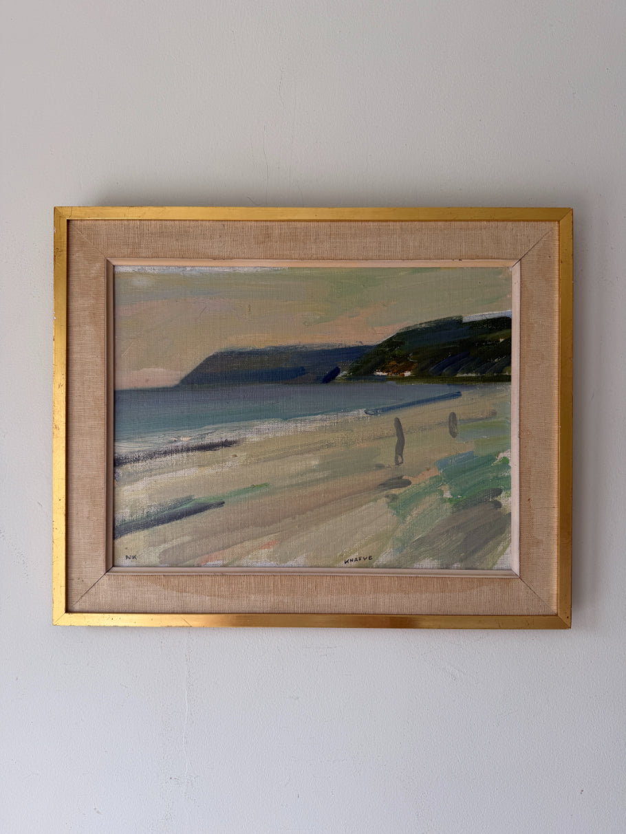 A framed vintage beach landscape painting with a gold frame. The painting depicts a sandy beach, ocean waves, dark cliffs, and a pale sky. Available at Hue. 