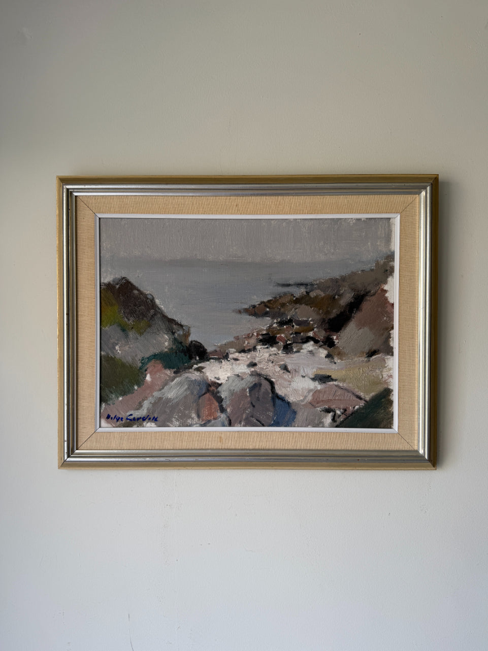 A framed vintage coastal landscape paintingl. The artwork depicts rocky cliffs, a shoreline, and muted water in soft gray, brown, white, and green tones. Available at Hue. 