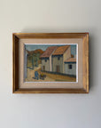 A framed vintage oil painting depicting a quiet village street with white buildings, terracotta roofs, and a palm tree. Available at Hue. 