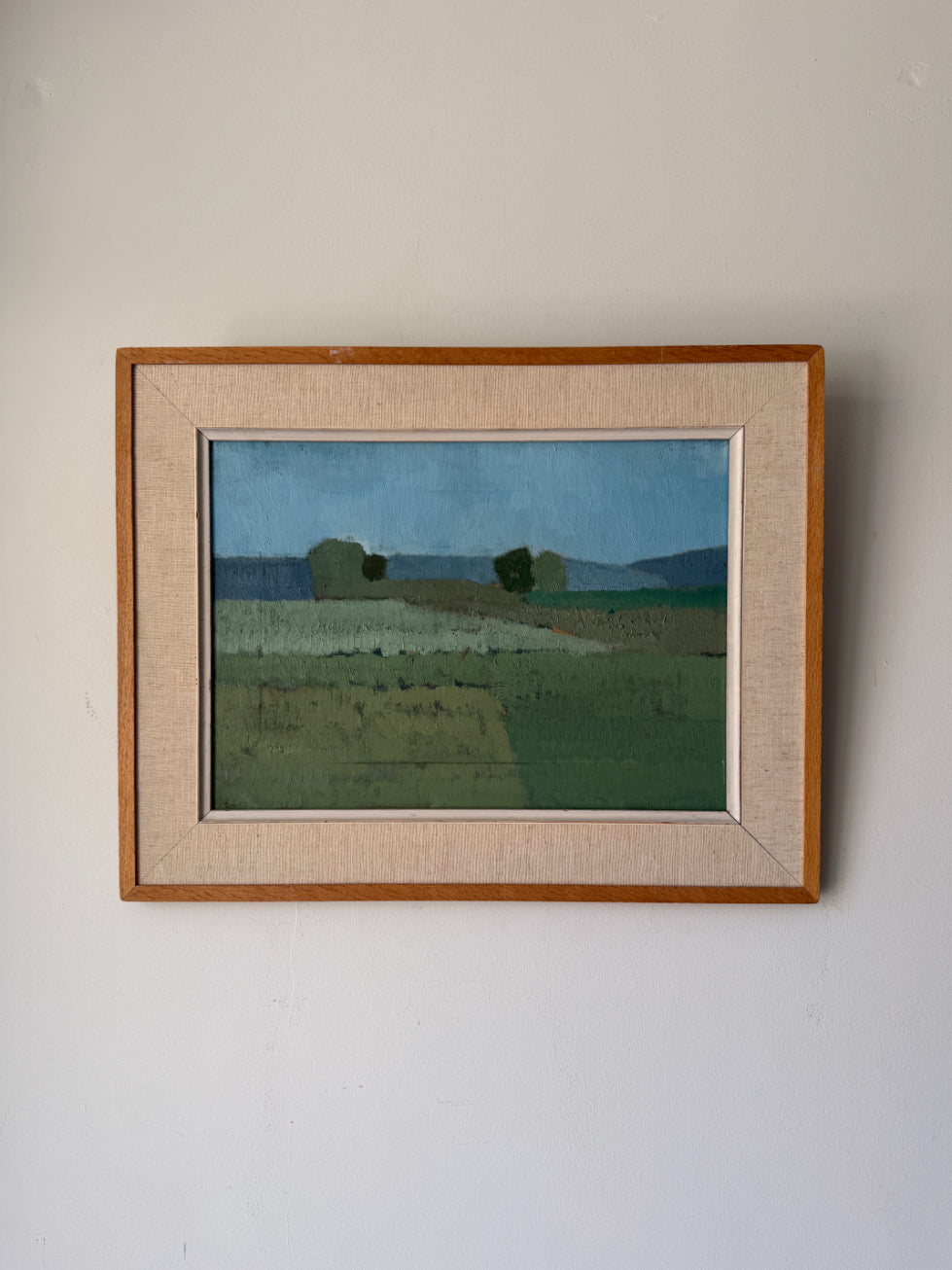 A framed vintage landscape oil painting featuring green fields and distant hills under a blue sky, housed in a light wood frame with a beige linen mat. Available at Hue.