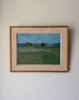 A framed vintage landscape oil painting featuring green fields and distant hills under a blue sky, housed in a light wood frame with a beige linen mat. Available at Hue.