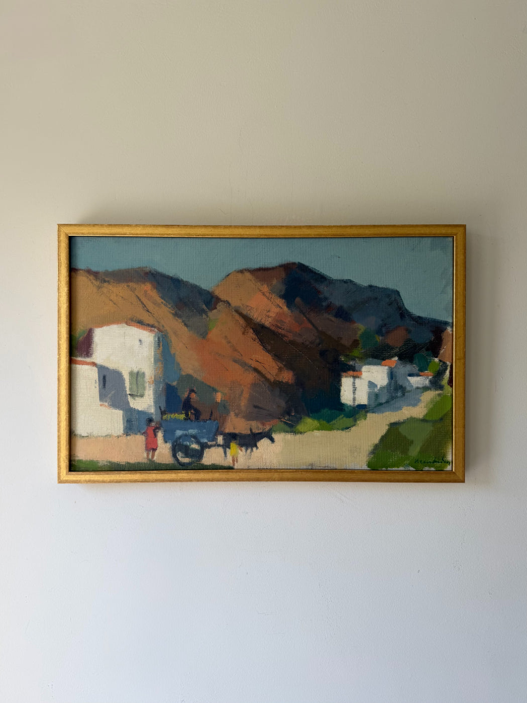 A framed vintage landscape oil painting depicting a village scene with white buildings, a horse-drawn cart, and brown mountains under a blue sky. Available at Hue. 