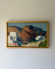 A framed vintage landscape oil painting depicting a village scene with white buildings, a horse-drawn cart, and brown mountains under a blue sky. Available at Hue. 