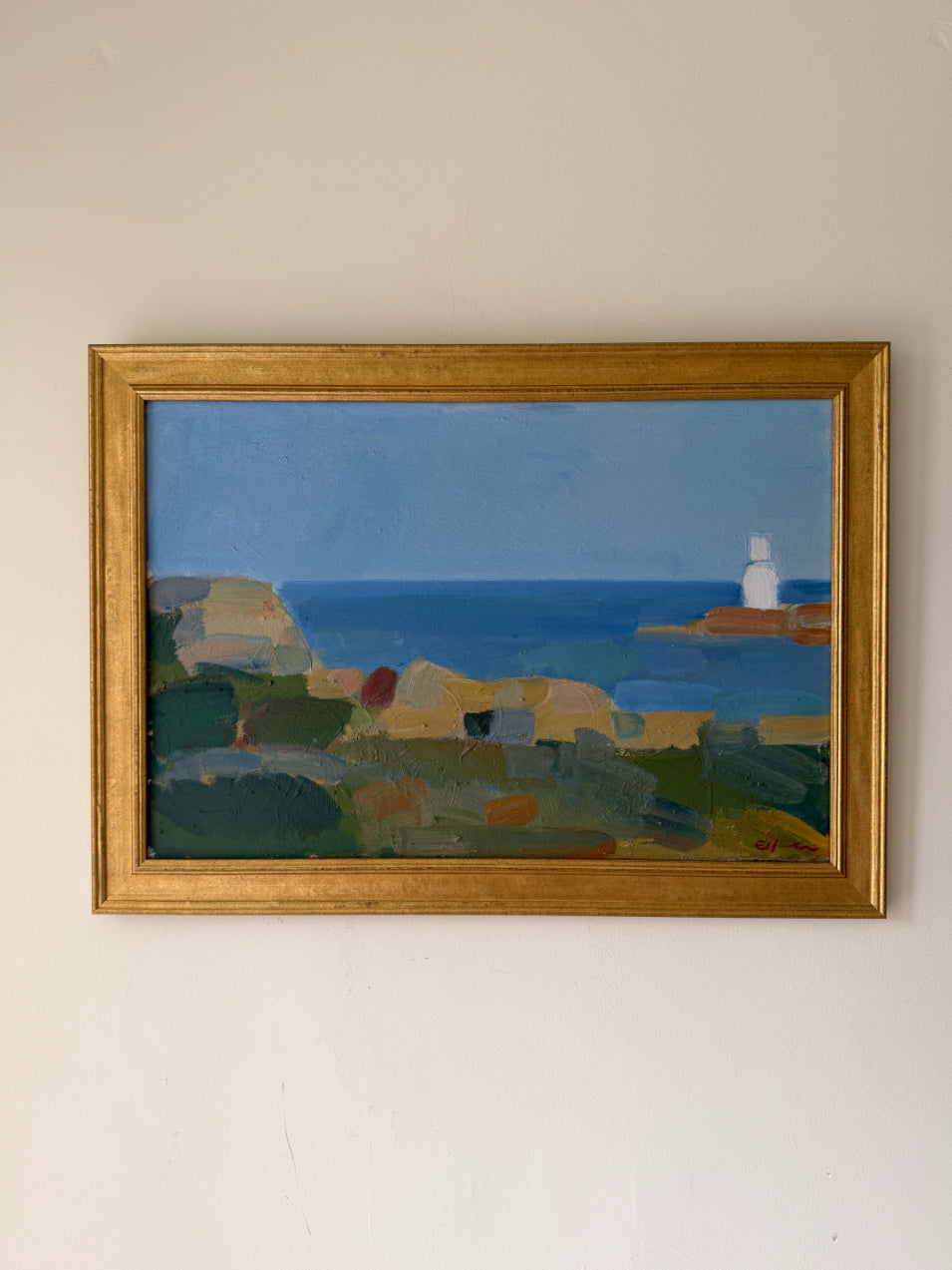A framed vintage landscape painting of a coastal scene with blue sea, green and beige cliffs, and a white lighthouse, housed in a gold frame. Available at Hue. 