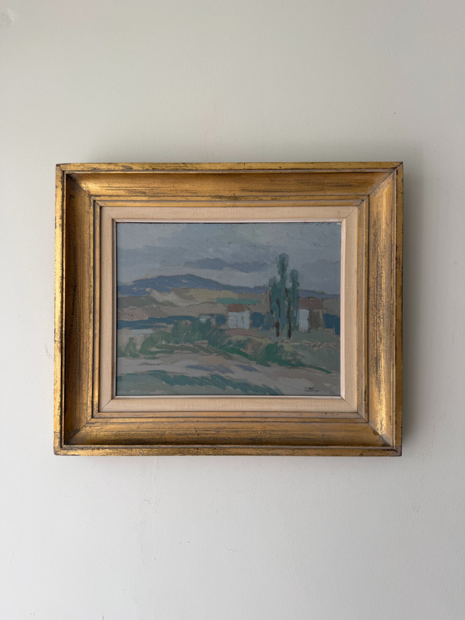 A framed vintage landscape oil painting. The artwork depicts a muted countryside scene with trees, hills, and small buildings in soft blue, green, and brown tones, enclosed in an ornate distressed gold frame. Available at Hue. 