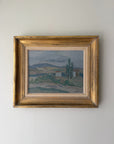 A framed vintage landscape oil painting. The artwork depicts a muted countryside scene with trees, hills, and small buildings in soft blue, green, and brown tones, enclosed in an ornate distressed gold frame. Available at Hue. 