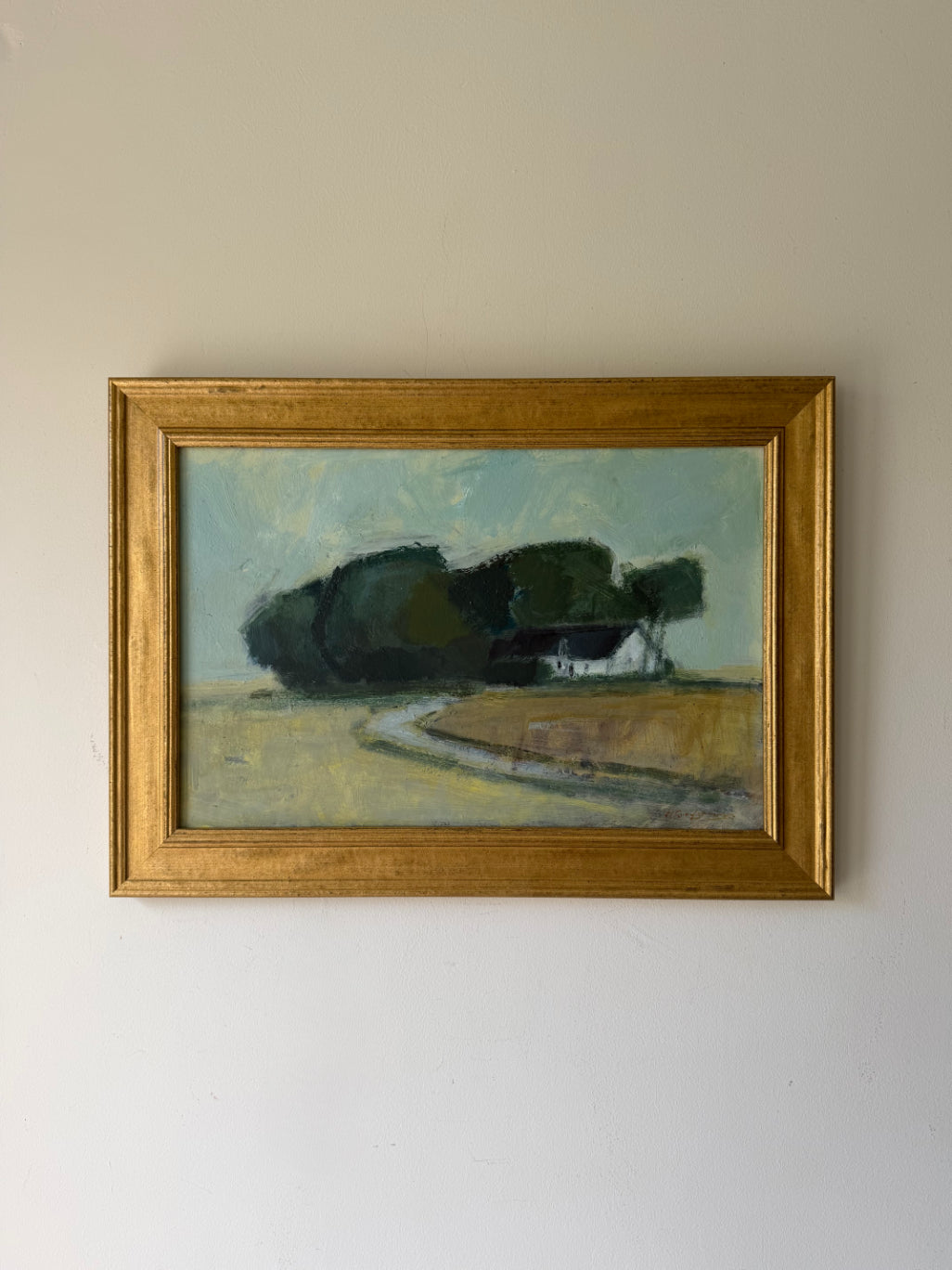 A framed vintage landscape oil painting featuring dark green trees, a small white house, and a winding road, housed in a gold frame. Available at Hue. 