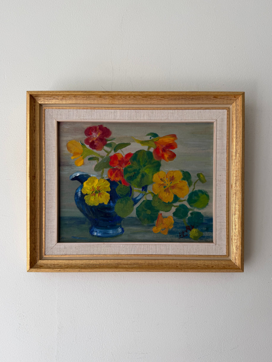 A framed vintage still life painting of colorful flowers in a blue pitcher vase The painting features yellow, red, and orange blossoms with green leaves, set against a muted blue-gray background, enclosed in a light wood frame with a beige mat.