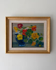A framed vintage still life painting of colorful flowers in a blue pitcher vase The painting features yellow, red, and orange blossoms with green leaves, set against a muted blue-gray background, enclosed in a light wood frame with a beige mat.