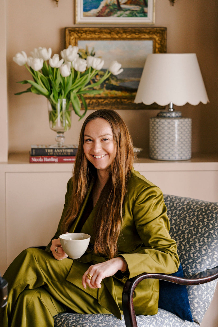 Art dealer and curator seated in a layered interior with vintage oil paintings, fresh flowers and decorative lighting, showcasing the personal approach behind Hue + Holasz.