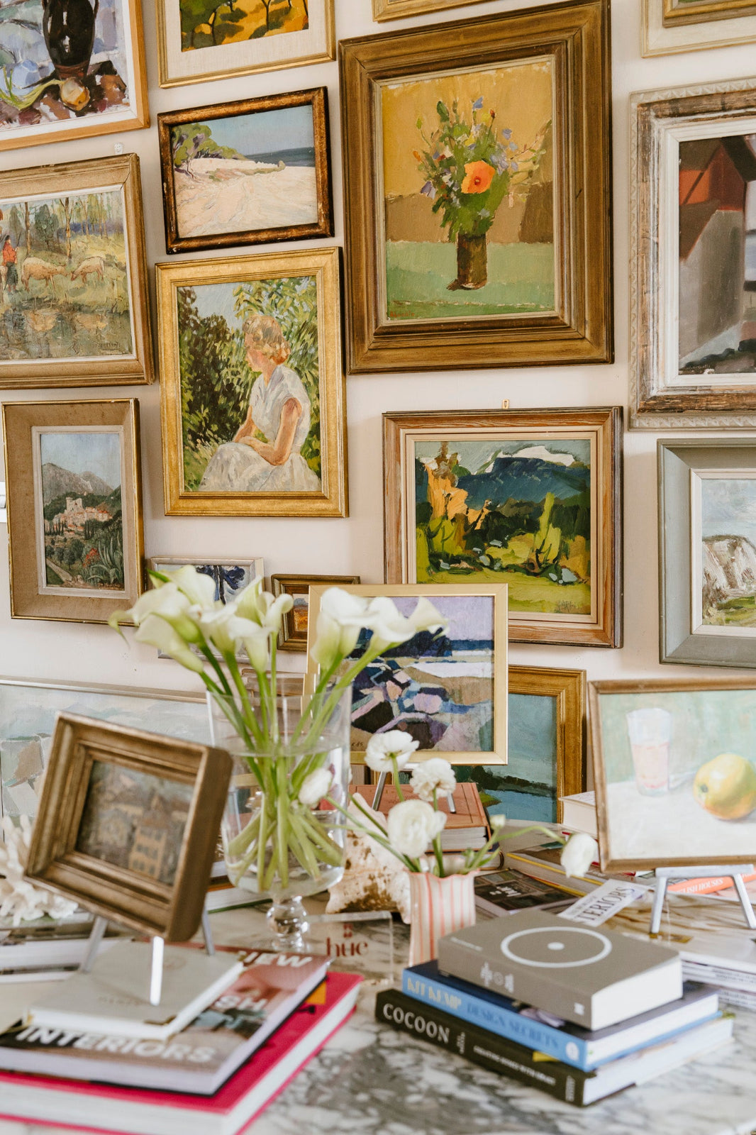 Eclectic gallery wall of framed vintage oil paintings and original art above a styled table with flowers, books and decorative objects, showcasing a layered collected interior.