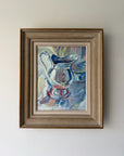 A framed vintage still life oil painting of a glass pitcher, featuring blue, white, and red brushstrokes, housed in a rustic brown wooden frame. Available at Hue. 